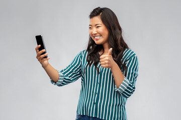 technology, blogging and people concept - happy asian woman taking selfie with smartphone or having video call and showing thumbs up over grey background