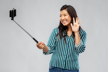 technology, blogging and people concept - happy asian woman taking picture with smartphone on selfie stick and waving hand over grey background