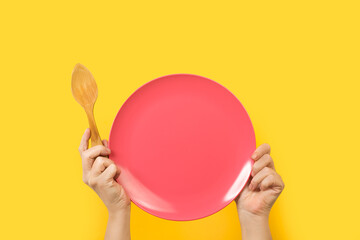 Manos sosteniendo un plato rosa vacío  y una cuchara de madera sobre un fondo amarillo liso y...