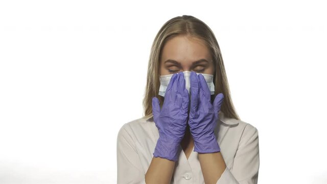 Tired nurse in robe smooths her face in mask with gloves. Blonde beautiful young woman looks at white background staring directly into camera. High quality 4k footage