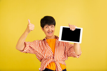 Shows blank tablet screen. Portrait of senior woman in stylish outfit isolated on yellow studio background. Tech and joyful elderly lifestyle concept. Trendy colors, forever youth. Copyspace for ad.