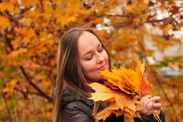 Naklejka na ściany i meble A smiling young woman holds yellow maple leaves in her hands. Happy young woman in autumn Park. A woman enthusiastically catches falling leaves. 