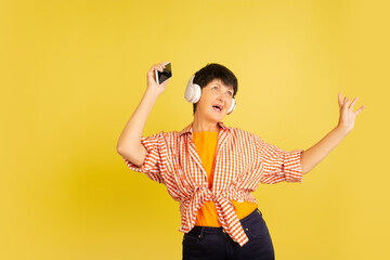 Listening to music with headphones. Portrait of senior woman in stylish outfit on yellow studio...