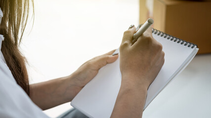 The hand of a business woman holds a pen and is taking information on a notebook with a blurred background.