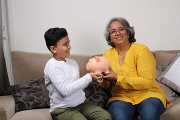 Indian/asian Grandma teaching her grand son importance of savings, putting coins into pink piggy bank.