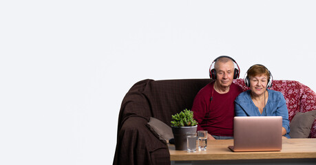 Fototapeta premium Banner. Senior husband and wife listen to music and talk over video communication using a computer with headphones at home. Photo with empty side space. Senior online and pandemic concept