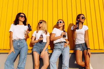 Obraz premium Smiling stylish young women in sunglasses with drinks standing against yellow wall in city on sunny summer day