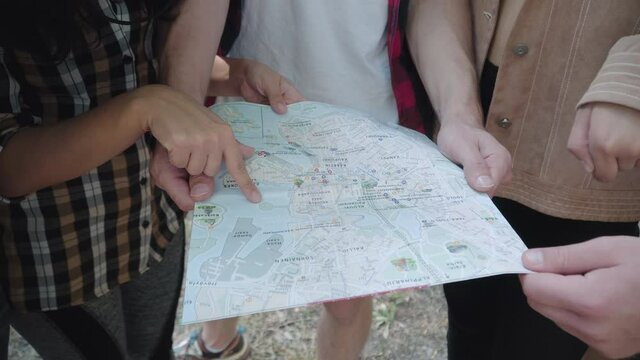 Close-up of paper map in male hands with unrecognizable men and women pointing and gesturing. Group of young Caucasian tourists hiking on sunny summer day outdoors. Eco-tourism concept.