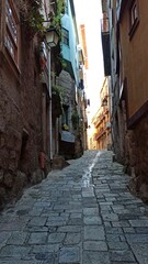 An ancient narrow street of an old European city. There is a very small distance between the colorful houses. The sidewalk is paved with large stone tiles. Laundry is dried on small balconies.
