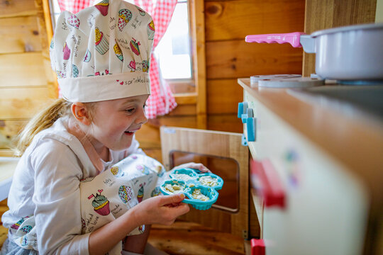 Young girl in wendy house putting cup cakes in oven pretending to cook in kitchen
