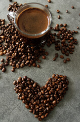 Cup of black coffee and coffee beans on grey table.  Heart shape on textured background. 