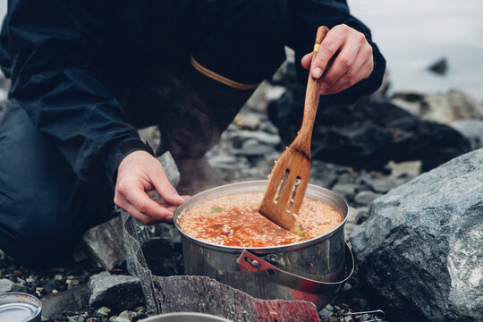 A wild camper stirring hot food in a pot cooking over a fire.