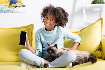 African american girl holding cellphone with blank screen, while stroking cat, sitting on sofa on blurred background