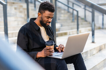 Young attractive Afro American businessman sitting on stairs in front of huge modern building and doing some work on laptop