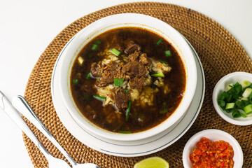Rawon, Indonesian traditional beef soup from Java Island. Served on white ceramic bowl topped with onion cracker and spring onion,chili paste and lime slices on rattan matt. Isolated white background.