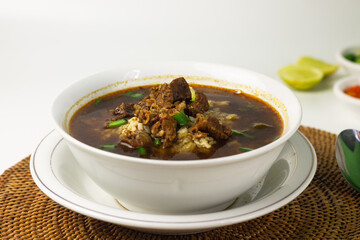 Rawon, Indonesian traditional beef soup from Java Island. Served on white ceramic bowl topped with onion cracker and spring onion,chili paste and lime slices on rattan matt. Isolated white background.