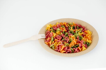 Multicolored pasta in a disposable paper plate with a wooden fork on a white background. Top view. Copy, empty space for text