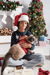 happy african american girl smiling at camera while cuddling cat near christmas tree on blurred background