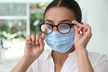 Woman wiping foggy glasses caused by wearing medical mask indoors, closeup