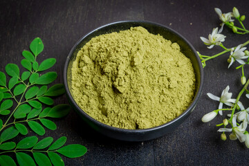 Moringa powder in black bowl ceramic, fresh green leaves and Moringa flowers on wooden background.