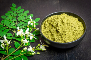 Moringa powder in black bowl ceramic, fresh green leaves and Moringa flowers on wooden background.