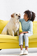 Smiling african american girl embracing and looking at retriever, while sitting on yellow sofa at home