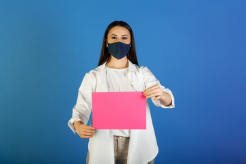 Pretty young woman in protective mask holding a pink blank sheet for your advertisement. Colourful studio portrait with blue background.