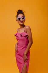 Confident african woman in long dress standing in studio. Elegant black model in pink outfit posing on yellow background.
