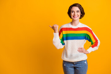 Photo of funny pretty brunette hair girl stand pointing look empty space wear rainbow sweater jeans isolated on bright yellow background