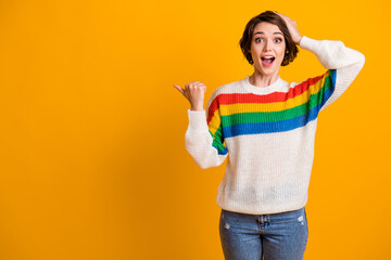 Photo of funny surprised girl open mouth stand pointing empty space hand head wear rainbow sweater blue jeans isolated on yellow background