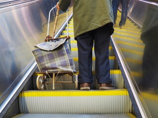 Elderly poor retired pensioner man standing with shopping cart and stick on subway escalator in...