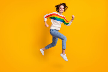 Fototapeta na wymiar Profile full size photo of cheerful crazy girl jump in air wear rainbow sweater denim jeans white shoes isolated on yellow background
