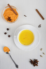 Top view of cup of golden or turmeric milk which is drink used as alternative remedy to boost immunity served on plate with spoon of curcuma powder and cinnamon on white wooden background. Vertical