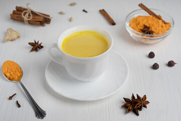 Cup of golden or turmeric milk which is Indian drink used as alternative remedy to boost immunity served on plate with spoon of curcuma powder and cinnamon on white wooden background. Horizontal image