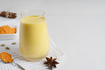 Glass of golden or turmeric milk which is Indian drink used as alternative remedy to boost immunity  served with spoon of curcuma powder and anise on white wooden background. Image with copy space