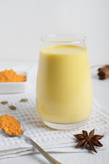 Glass of golden or turmeric milk which is Indian drink used as alternative remedy to boost immunity served on textile towel with spoon of curcuma powder and anise on white wooden background. Vertical