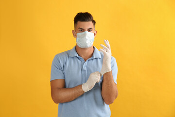 Man in protective face mask putting on medical gloves against yellow background