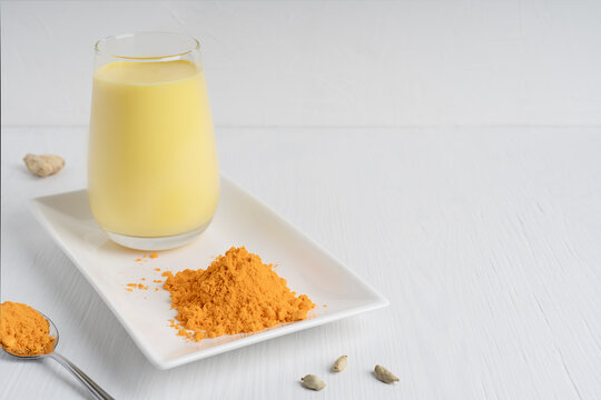 Glass of golden or turmeric milk which is Indian drink used as alternative remedy to boost immunity served on plate with spoon of curcuma powder on white wooden background. Image with copy space