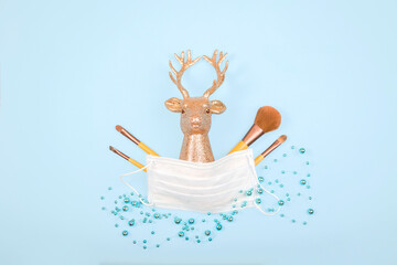 Golden deer head with medical mask, cosmetics brushes on blue background with New Year balls, lights. Xmas holidays in new Covid-19 reality.