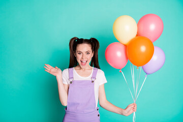 Photo of lovely young girl hold balloons waving palm open mouth wear purple overall isolated teal color background