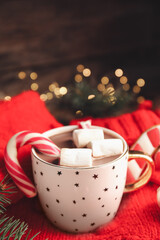 Cup of tasty cocoa with marshmallows and Christmas candy cane on knitted sweater against blurred festive lights. Space for text