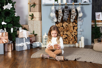 A cute little girl in a white sweater sits near the fireplace and an elegant Christmas tree and holds a brown gift, sadly looks at the gift