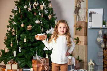 A cute little girl in a white sweater stands next to an elegant Christmas green christmas tree and holds an eco gift, looks at the camera and smiles