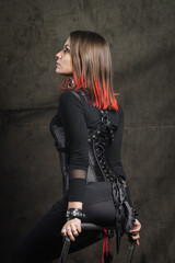Fototapeta na wymiar A attractive dominant woman with piercings and bright hair in a black corset, with leather harnesses and bracelets posing against the background of an old black wall.
