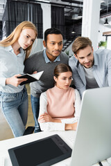 Multicultural businesspeople with notebook looking at computer near graphics tablet on blurred foreground in office