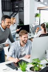 Smiling multiethnic colleagues using graphics tablet and computer near plants on blurred foreground in office