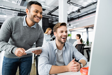Cheerful multiethnic businessmen with digital tablet looking at computer monitor on blurred foreground in office