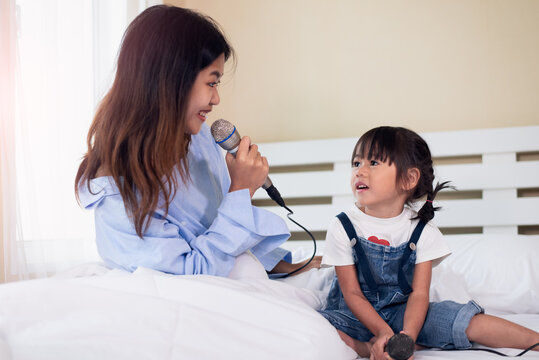 Happy Asian Family Loving Children, Kid And Her Sister Holding Microphone And Singing Together On Bed In Bedroom