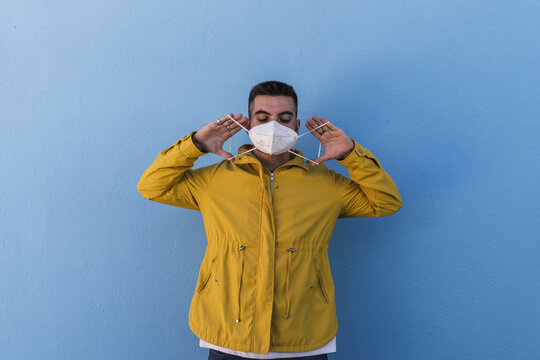 Young male wearing a yellow jacket and putting on a facemask against a wall - COVID-19
