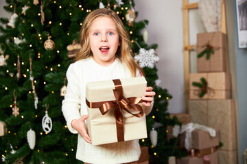 The girl stands in a room with a green Christmas tree and holds a gift, the girl received a gift for Christmas and happy, the girl looks into the camera